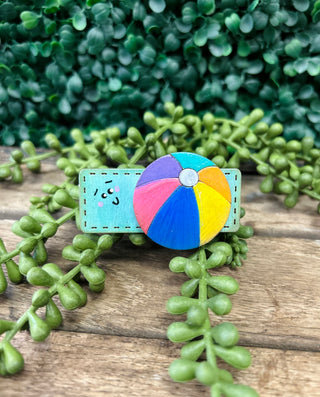 Colorful handmade kids hair clip with cute character design displayed on wooden surface with green plants.