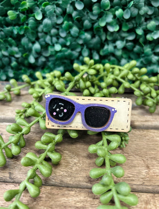 Handmade kids hair clip with purple sunglasses design and cute face on wooden surface with green plants.