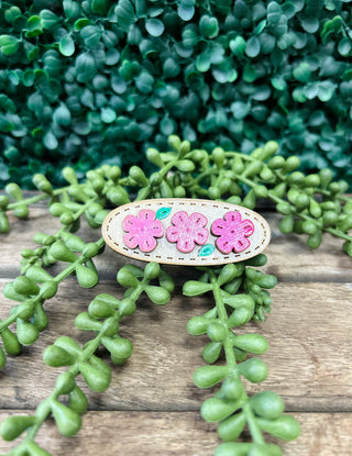 Handmade oval hair clip with embroidered pink flowers on wooden surface surrounded by green succulent plants.