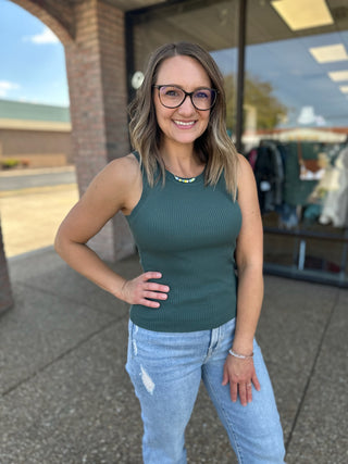 Woman wearing green knit ribbed tank top with jeans, smiling outdoors at boutique storefront.