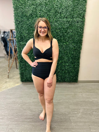 Woman wearing black two-piece bikini with high-waisted bottoms and wrap-style top, standing against green wall.