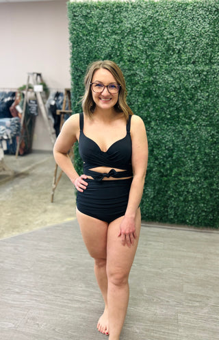Woman wearing black high-waisted two-piece bikini with cutout details, smiling in retail store setting.