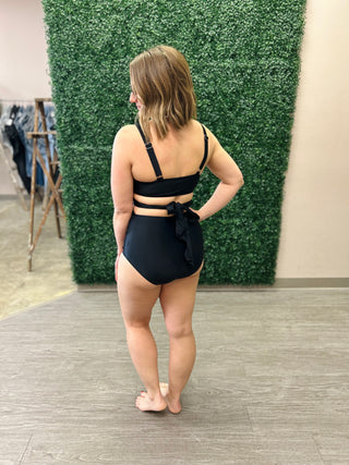 Woman modeling black high-waist bikini with strappy details from behind in retail setting.