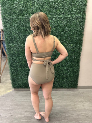 Woman modeling taupe high-waisted bikini with wrap-style top from behind against green hedge backdrop.