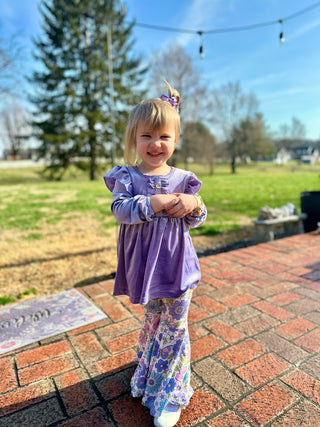 Young girl wearing purple top and floral pants outfit standing on brick patio outdoors