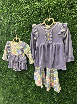 Two matching girls' outfits with purple ruffled tops and floral pants hanging on heart-shaped hangers against greenery.