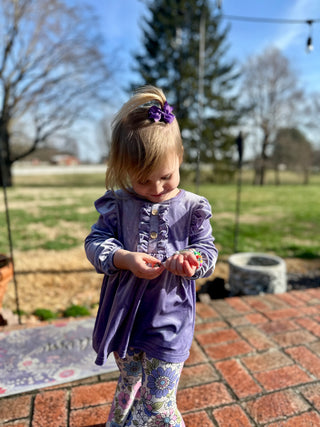 Young girl wearing purple ruffled top and floral leggings standing outdoors on brick patio
