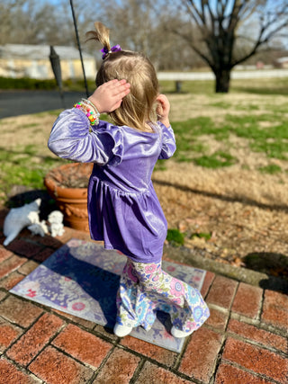 Young girl wearing purple velvet top and floral leggings outfit standing on brick patio outdoors.