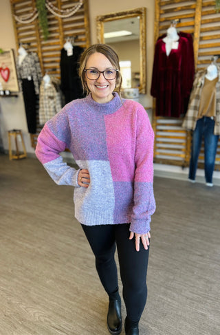 Woman wearing colorful block-patterned sweater standing in clothing boutique with fashion displays in background.