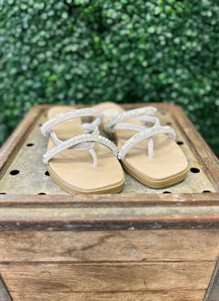 Ivory beaded flip-flop sandals with ankle straps displayed on rustic wooden crate against green foliage background.