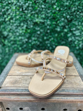 Gold beaded sandals with embellished straps displayed on wooden surface against green foliage background.
