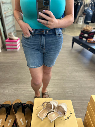 Woman wearing light wash denim high-rise shorts with double button waistband, taking mirror selfie in store.
