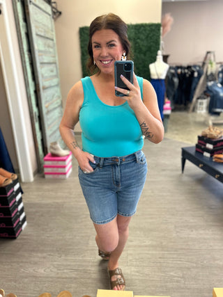 Woman wearing turquoise tank top and denim shorts taking mirror selfie in retail store
