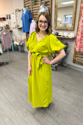 Woman wearing bright lime green maxi dress with flutter sleeves and belt in clothing boutique store.