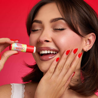 Woman with red nails holding Poppy & Pout Smarties Cherry Vanilla lip balm against red background