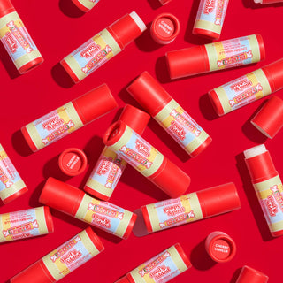 Multiple red and cream colored Smarties Cherry Vanilla lip balm tubes scattered on bright red background.
