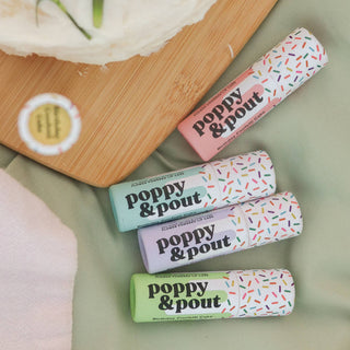 Four colorful Poppy & Pout lip balms with sprinkle design arranged on green surface with wooden cutting board.