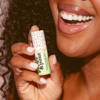 Woman with white manicured nails holding colorful sprinkle-patterned lip balm tube while smiling, showing white teeth.