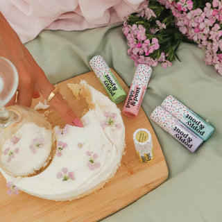 Lip balm tubes with sprinkle pattern design displayed alongside frosted cake and pink flowers on wooden board.