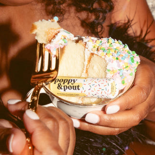 Hands holding birthday confetti cake lip balm with colorful sprinkles and gold fork decoration.