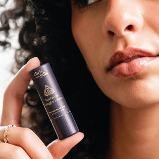 Person holding Chris Loves Julia Blackberry Campfire S'mores lip balm tube near their glossy lips.