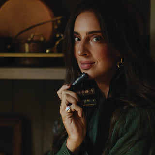 Woman holding dark lip balm tube in cozy indoor setting with warm lighting and green sweater.