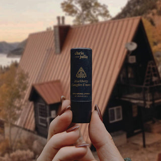 Hands holding Poppy & Pout lip balm tube outdoors with rustic cabin in blurred background.
