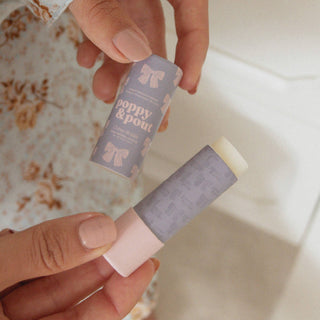 Hands holding Poppy & Pout lip balm tube and cap above bath water with towel visible