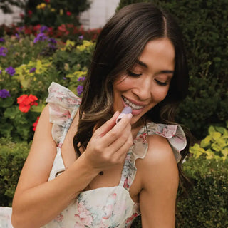 Woman in floral dress applying lip balm outdoors with colorful garden flowers in background.