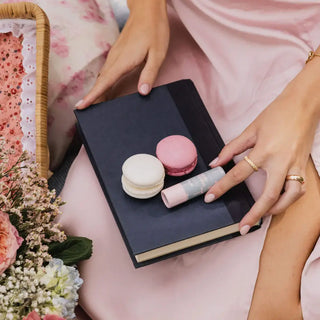 Hands holding French macaron-shaped lip balms on black book with pink floral styling and basket.