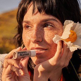 Woman holding Poppy & Pout lip balm while posing with a white flower near her face.