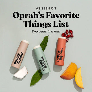 Poppy & Pout lip balm tubes in various shades surrounded by mint leaves, fruit slices, and pomegranate seeds.