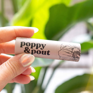 Hand holding Poppy & Pout lip balm tube with floral design against green leafy background.