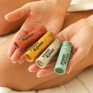 Hand holding four colorful Poppy & Pout lip balm tubes with floral botanical packaging designs.