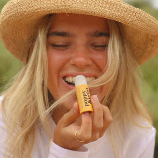 Young woman wearing straw hat smiling while applying Poppy & Pout lip balm outdoors in natural sunlight.