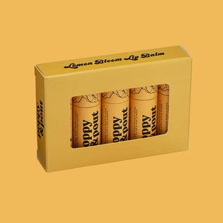 Yellow gift box containing three tubes of Poppy & Pout Lemon Bloom lip balm on orange background.