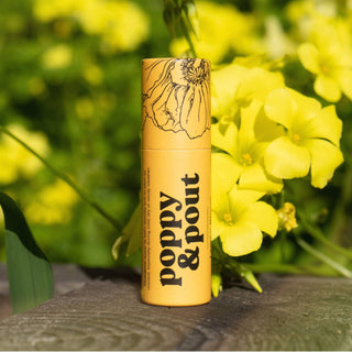 Poppy & Pout lip balm tube with floral design sitting on wooden surface surrounded by yellow flowers