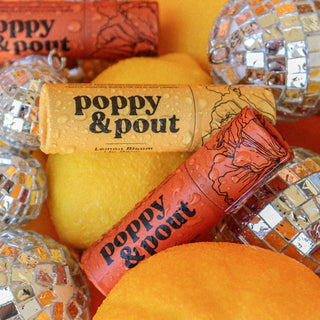 Poppy & Pout lip balm tubes displayed with decorative disco balls on orange background