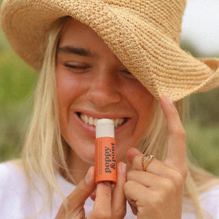 Woman in straw hat smiling while holding orange Poppy & Pout lip balm tube outdoors.