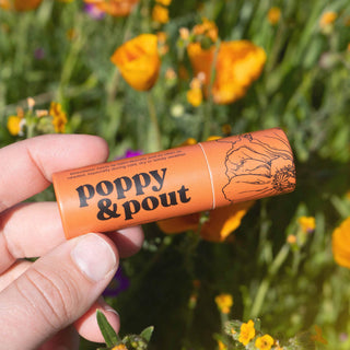 Hand holding orange Poppy & Pout lip balm tube with floral design against blooming wildflower background