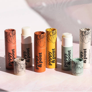 Collection of Poppy & Pout lip balms with floral packaging in various colors displayed on pink surface
