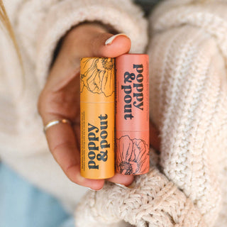 Hand holding two Poppy & Pout lip balm tubes with floral packaging against knitted sweater background.