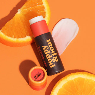 Poppy & Pout lip balm tube with orange slices on vibrant orange background in natural lighting.