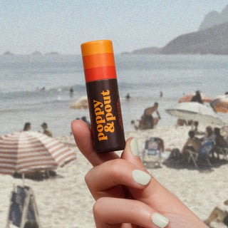 Hand holding Poppy & Pout lip balm tube on sandy beach with ocean and beachgoers in background.