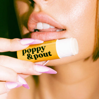 Woman with pink manicured nails holding Poppy & Pout lip balm tube near her glossy lips.