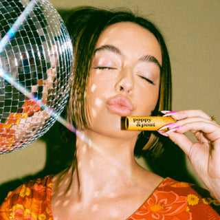Woman with closed eyes holding Poppy & Pout lip balm next to disco ball in retro setting.
