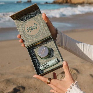 Person holding open Poppy & Pout lip care duo gift box on beach with ocean background