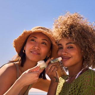Two women sharing Poppy & Pout Sweet Mint lip balm outdoors under blue sky