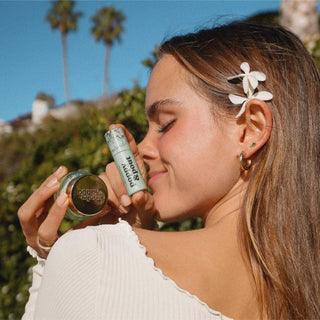 Woman holding Poppy & Pout lip balm and lip scrub products outdoors with white flowers in hair.