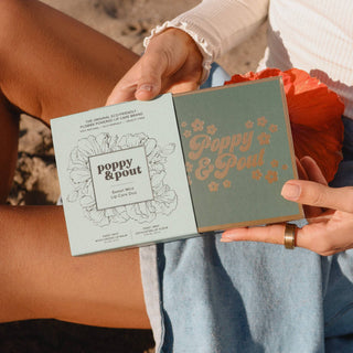 Hands holding Poppy & Pout lip care duo package with floral design and product information visible.